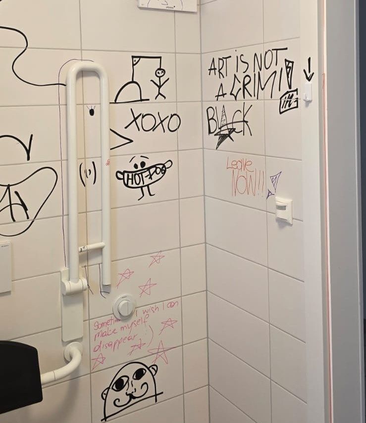 Eine Badezimmerwand mit Graffiti und Nachrichten. Es enthält Zeichnungen von Gesichtern, einer Person und einem Hund, mit Phrasen wie 'Kunst ist kein Verbrechen', 'xoxo', 'Schwarz' und 'Leggie'. Es gibt auch einen Handlauf und ein weißes Objekt an der Wand.