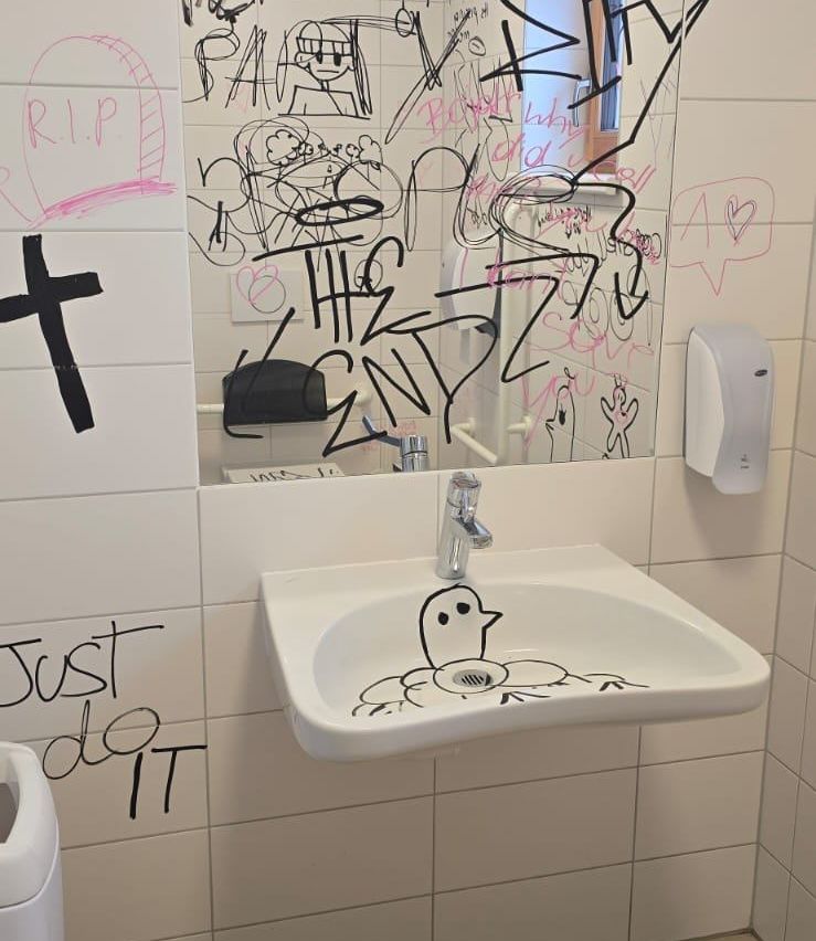 Ein Badezimmer mit einem Waschbecken, das eine Geisterzeichnung zeigt, umgeben von Graffiti an den Wänden und Fliesen, einschließlich eines Kreuzes und eines Herzens mit RIP.