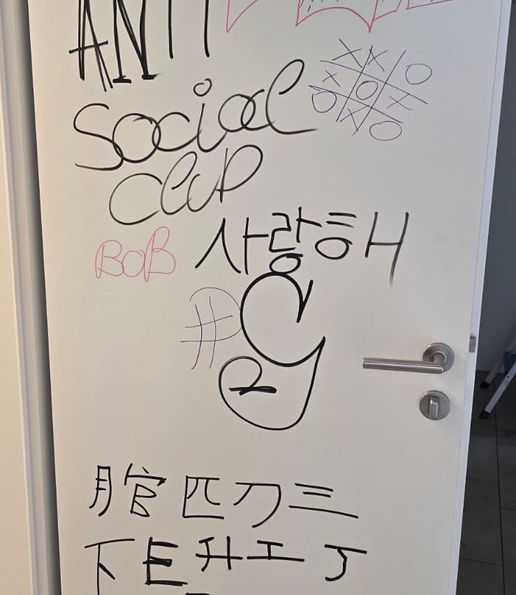 Eine weiße Tür mit Graffiti, darunter 'ANTI Social Club', ein Name 'Bob', Symbole und fremdsprachiger Text. Es gibt zwei silberne Griffe an der Tür.