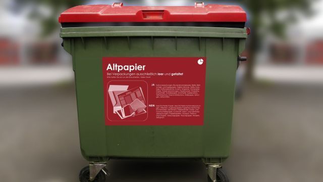A green waste bin with a red lid and a label reading 'Altpapier'. The label includes a symbol and text about recycling old paper. The bin is on a road with blurred trees and buildings in the background.