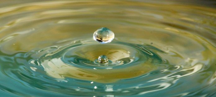 Eine Wassertropfen schwebt über einem Teich und erzeugt Wellen, während er das Wasser sanft berührt. Der Tropfen reflektiert das Licht und fügt einen schimmernden Effekt hinzu.
