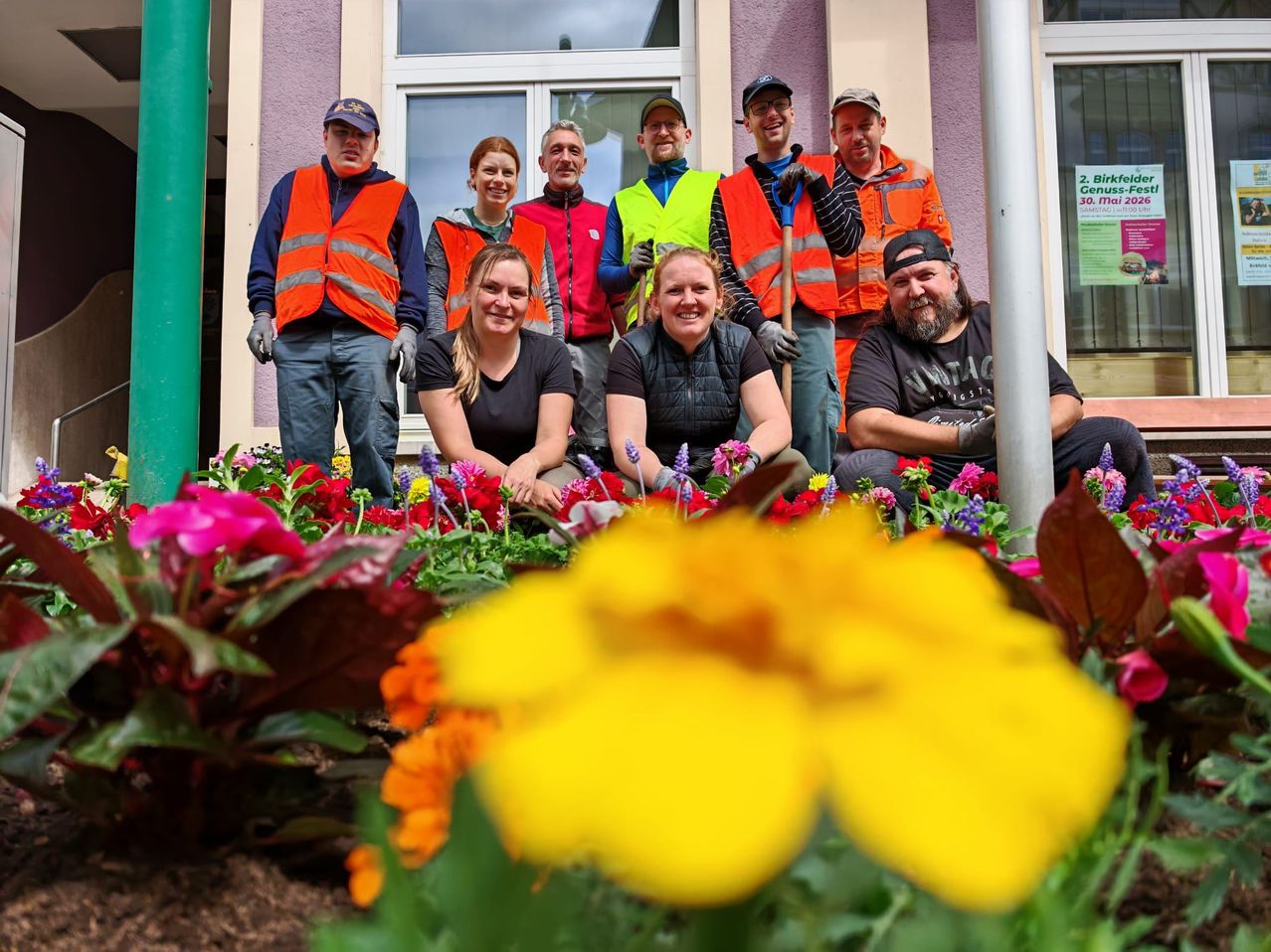 Eine Gruppe von Freiwilligen in Sicherheitswesten posiert für ein Foto in einem Garten, mit Blumen und Pflanzen im Vordergrund.