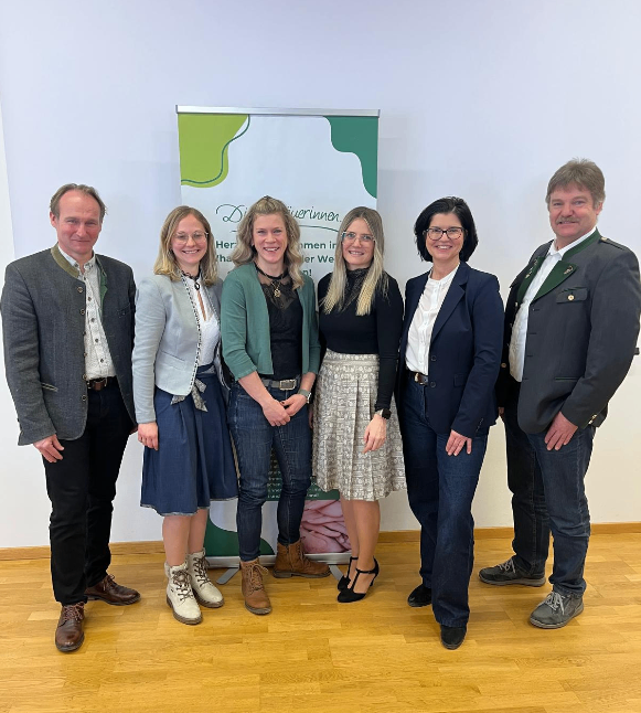 Six people, three women and three men, stand in front of a banner, smiling for a photo. The men wear suits and leather shoes, and the women wear skirts, glasses, and boots. The banner has green and white colors with text.