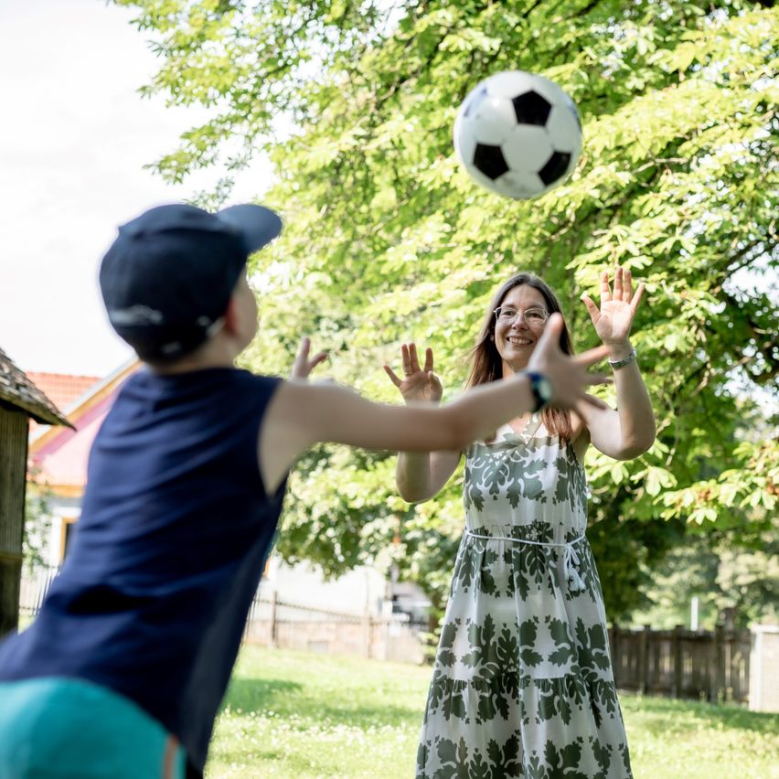 Eine Frau und ein Kind spielen in einem Park mit einem Fußball. Das Kind streckt die Hand nach dem Ball aus, während die Frau lächelt und zusieht.