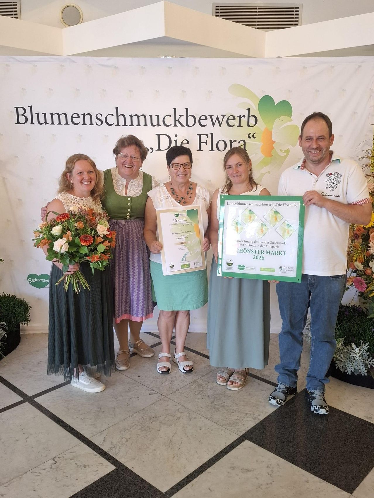 Fünf Personen stehen vor einem Banner mit dem Text 'Blumenschmuckbewerb, Die Flora'. Sie lächeln und halten Urkunden und einen Blumenstrauß. Der Mann rechts trägt ein weißes Poloshirt mit Logo.