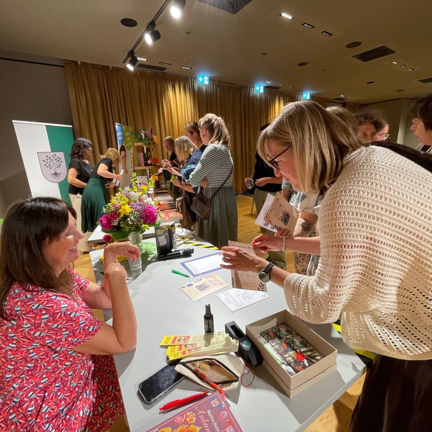 Several women are seated around a table with various items, including flowers, books, and a phone. One woman reads a paper while another holds a box. A sign with a logo is in the background.