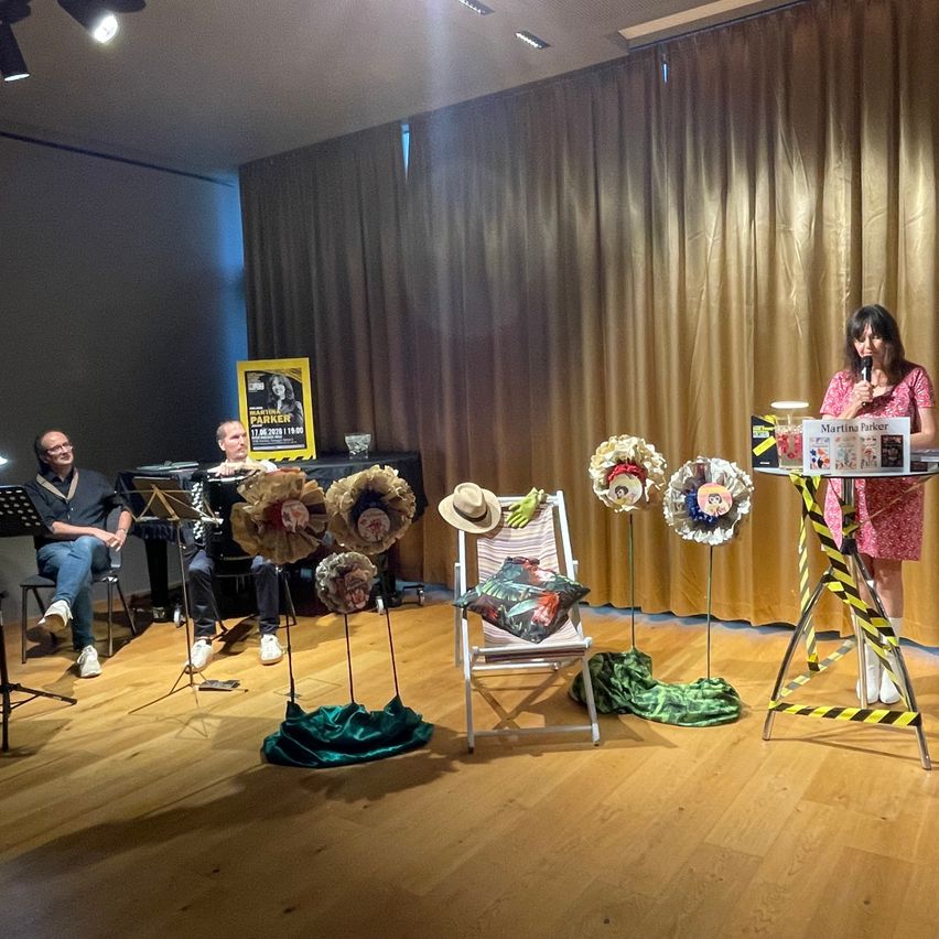 A woman is speaking into a microphone on a stage, with a man playing the piano behind her and another man sitting. There are decorative flower displays and a table with a book.