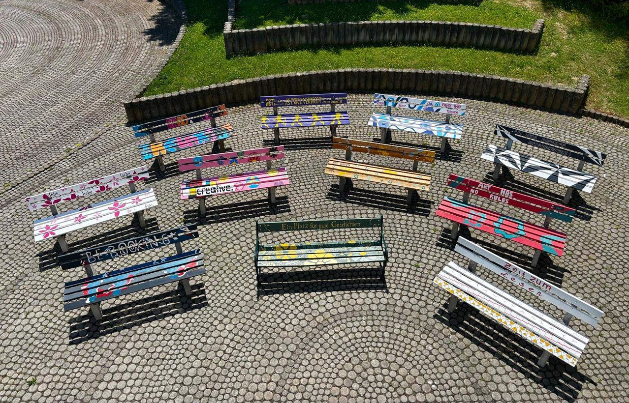 An aerial view of a park with benches painted in various colors and designs arranged in a circle. The benches are on a cobblestone floor.