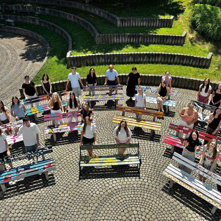 A group of people stand and sit around decorated benches in an outdoor amphitheater. Some benches have German writing. The area is surrounded by steps and greenery.