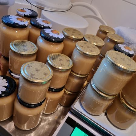 Mehrere Gläser Honig mit weißen und gelben Gänseblümchendeckeln, gestapelt auf einer Metalloberfläche und einer digitalen Waage. Die Honiggläser sind auf einer Edelstahloberfläche.