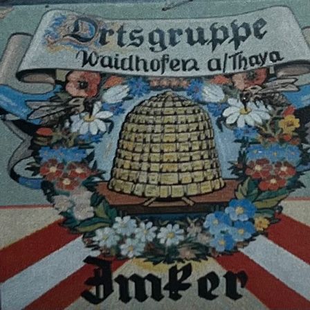Das Bild zeigt ein dekoratives Emblem mit einem Bienenstock in der Mitte, umgeben von einer Blumengirlande. Das Emblem befindet sich auf einem rot-weiß gestreiften Hintergrund, mit einem Banner darüber, das Ortsgruppe Waidhofen a/Thaya liest. Unten steht das Wort Imker.