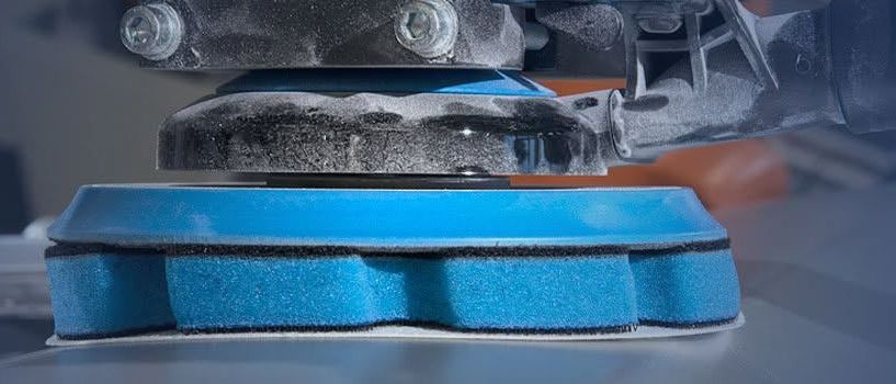 Eine Nahaufnahme eines blauen und grauen industriellen Schleifers mit Metallrahmen und blauem Schleifscheiben. Die Maschine ist auf einer grauen Oberfläche positioniert.