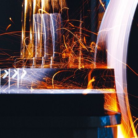 Eine Nahaufnahme einer Metallbearbeitungsmaschine mit Funkenflug aus dem Schneidprozess. Die Maschine ist von einem hellen Licht beleuchtet, das Schatten auf die Metallteile wirft.