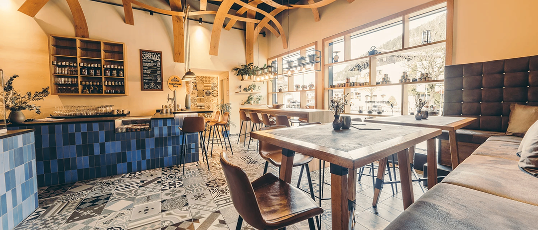 Ein Coffee-Shop-Interieur mit Holzdecke, gefliestem Boden und Holztischen. Die Tische haben Lederstühle und dekorative Vasen. Die Wände sind mit Regalen und einer Tafel dekoriert. Der Raum hat große Fenster und ist gut beleuchtet.