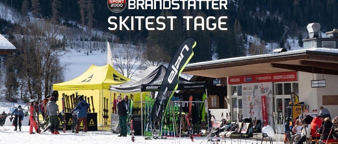 Ein Skitest-Event findet in einem schneebedeckten Außenbereich statt, mit Menschen, Zelten und Skiausrüstung. Im Hintergrund befinden sich mit Schnee bedeckte Berge.