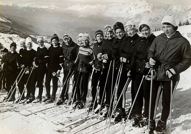 Eine Gruppe von Menschen in Winterkleidung steht in einer Reihe auf einem verschneiten Berg. Sie halten Skistöcke und tragen Skischuhe.
