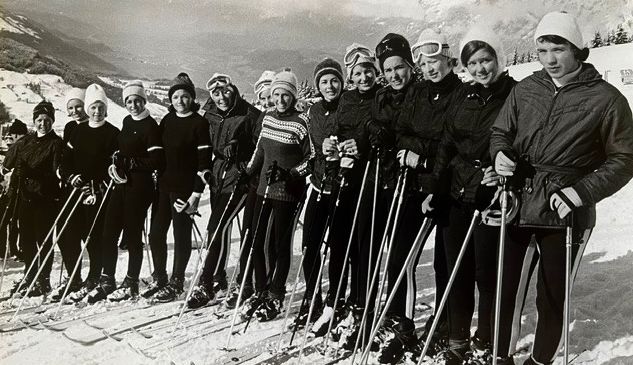 Eine Gruppe von Menschen in Winterkleidung steht in einer Reihe auf einem verschneiten Berg. Sie halten Skistöcke und tragen Skischuhe.