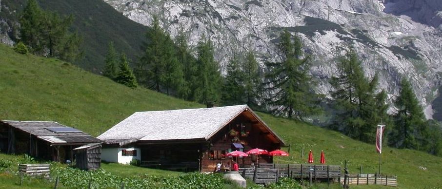 Ein kleines Chalet in einem Berggebiet mit einigen Leuten, die unter roten Sonnenschirmen sitzen.