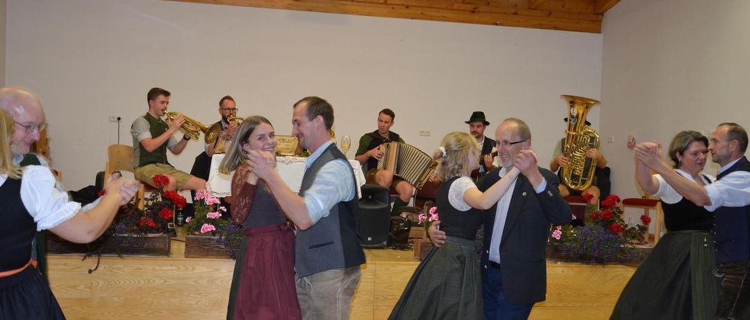 Eine Gruppe von Menschen tanzt auf einer Bühne, während eine Band im Hintergrund spielt. Der Mann und die Frau vorne lächeln und halten Händchen. Dahinter spielt ein Mann Trompete und ein anderer Akkordeon. Es gibt Blumen und einen Tisch mit einer Flasche auf der Bühne.