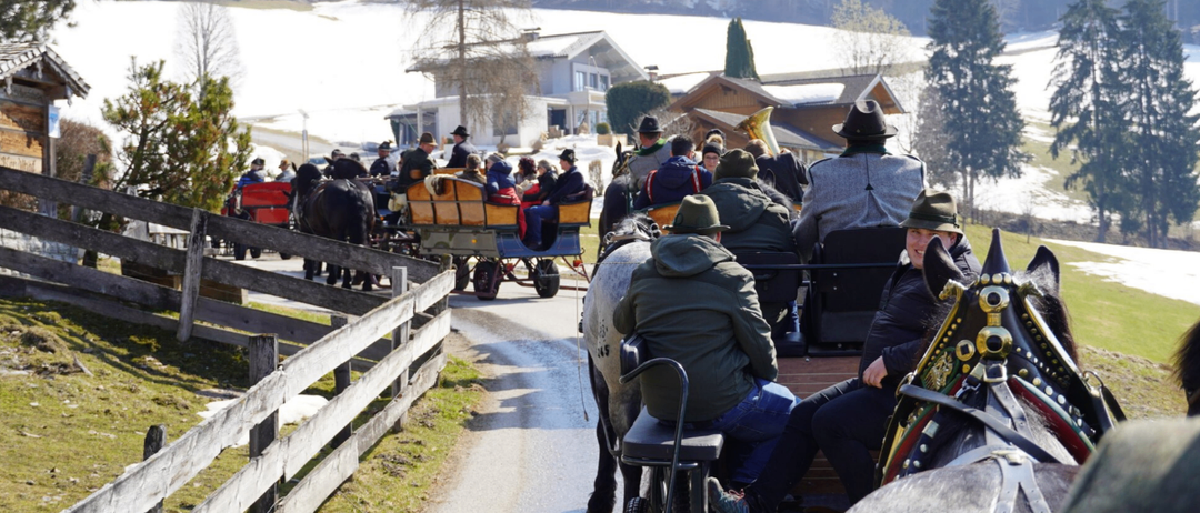 Eine Gruppe von Menschen fährt auf von Pferden gezogenen Kutschen in einer verschneiten Landschaft. Sie sind warm gekleidet und einige spielen Musikinstrumente. Die Kutschen bewegen sich auf einer Straße, umgeben von Häusern und Bäumen.