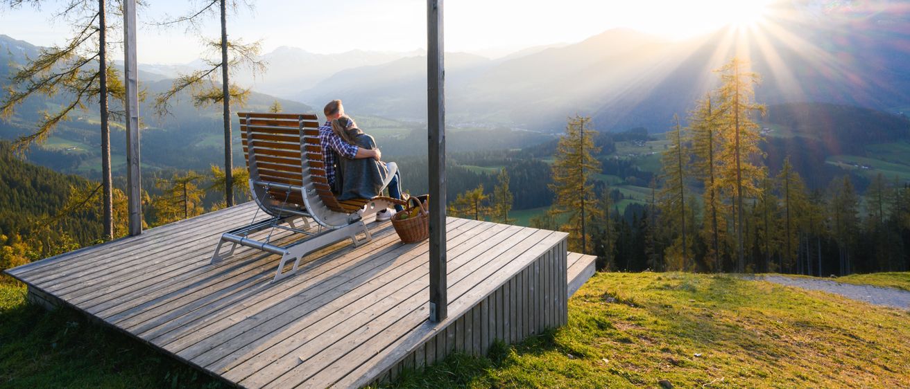 Ein Paar sitzt auf einer Bank auf einer Holzterrasse und umarmt sich, während es den malerischen Bergblick bei Sonnenaufgang genießt.