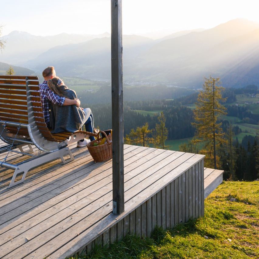 Ein Paar sitzt auf einer Bank auf einer Holzterrasse und umarmt sich, während es den malerischen Bergblick bei Sonnenaufgang genießt.
