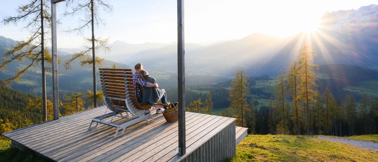 Ein Paar sitzt auf einer Bank auf einer Holzterrasse und umarmt sich, während es den malerischen Bergblick bei Sonnenaufgang genießt.