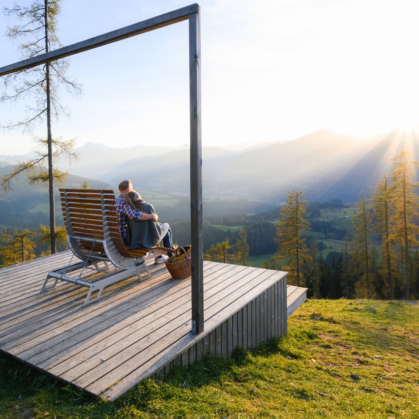 Ein Paar sitzt auf einer Bank auf einer Holzterrasse und umarmt sich, während es den malerischen Bergblick bei Sonnenaufgang genießt.