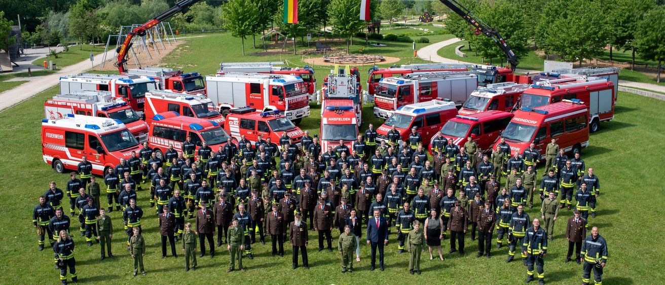 Eine große Gruppe von Feuerwehrleuten und Beamten steht vor vielen Feuerwehrfahrzeugen auf einem Grasfeld. Die Zahl 732 ist mit gelben Helmen gemacht.