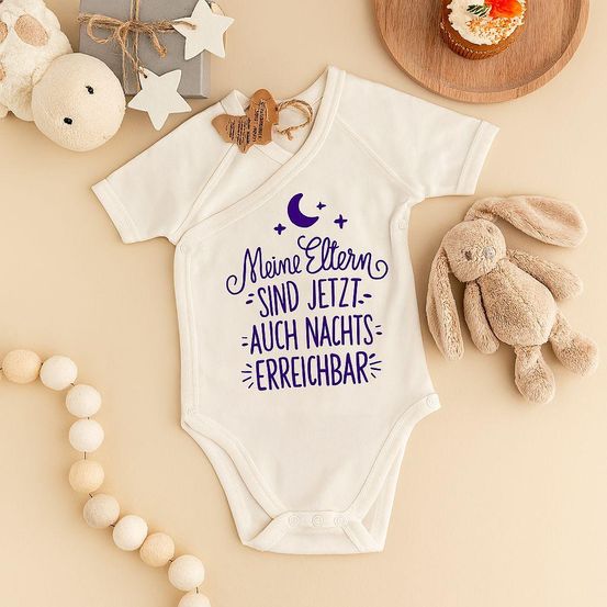 Ein weißer Baby-Bodysuit mit lila Text wird von einem Cupcake, Plüschtieren, einem Holzteller und dekorativen Sternen umgeben. Der Text lautet 'Meine Eltern sind jetzt auch nachts erreichbar.'
