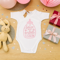Ein weißes Babybody mit pinkem Text liegt auf einem Holztisch. Der Text lautet 'Ich Schreie Nichti Ich Bestelle Essen'. Ein Plüschtier, Luftballons und Geschenke sind ebenfalls auf dem Tisch.