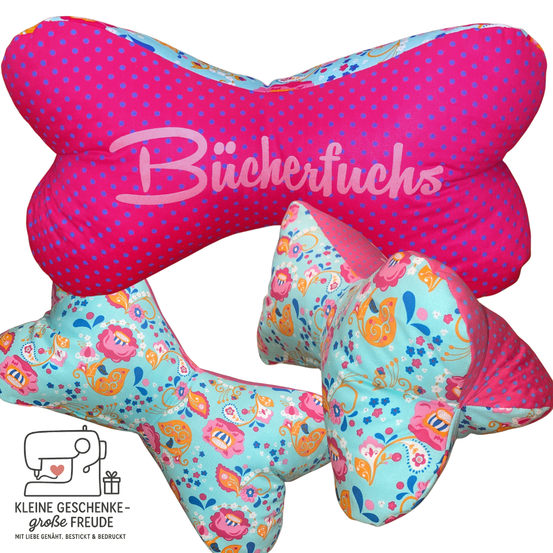 Ein rosafarbenes Hundekissen mit Punkten und dem Wort Buchertuschs in rosa Buchstaben. Ein türkisfarbenes und rosa Hundekissen mit Blumendesigns. Die Kissen stehen auf weißem Hintergrund mit einem Nähmaschinenlogo und einem Geschenk.
