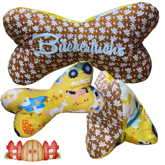 Drei knochenförmige Kissen mit verschiedenen Mustern. Das oberste hat das Wort Buchertüchs in Blau geschrieben. Das mittlere hat einen gelben Traktor und Bauernhoftiere. Das unterste hat eine gelbe Kuh und ein braun-weißes Muster.