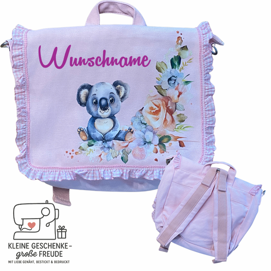 Ein lila Rucksack mit Koala-Bär-Illustration und Blumendesign, der den Text 'Wunnschname' zeigt. Ein kleinerer rosa Rucksack ist daran befestigt. Unten sind ein Nähmaschinenlogo und ein Geschenkkarton zu sehen.