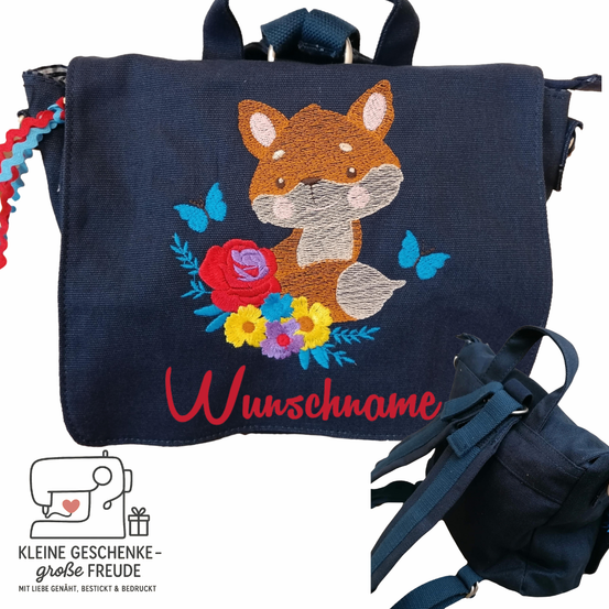 Ein blauer Rucksack zeigt einen gestickten Fuchs, umgeben von Blumen und Schmetterlingen. Der Name Wunschname ist rot gestickt. Im Vordergrund ist eine kleinere Tasche zu sehen.