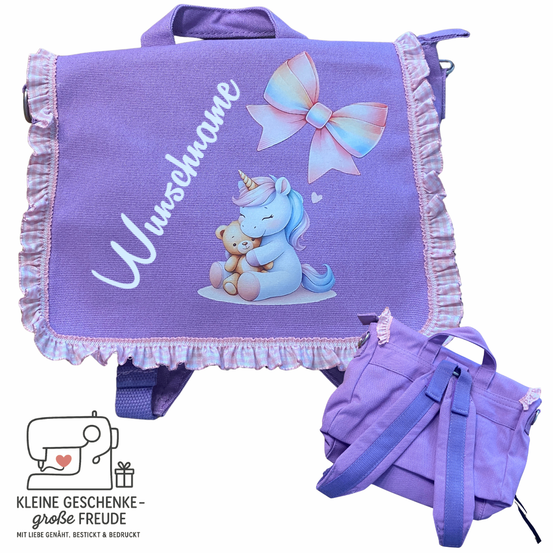 Ein lila Rucksack mit einem Einhorn und einem Teddybär. Der Rucksack ist mit einer Schleife verziert und hat Rüschen an den Kanten. Ein kleinerer Beutel ist ebenfalls zu sehen.