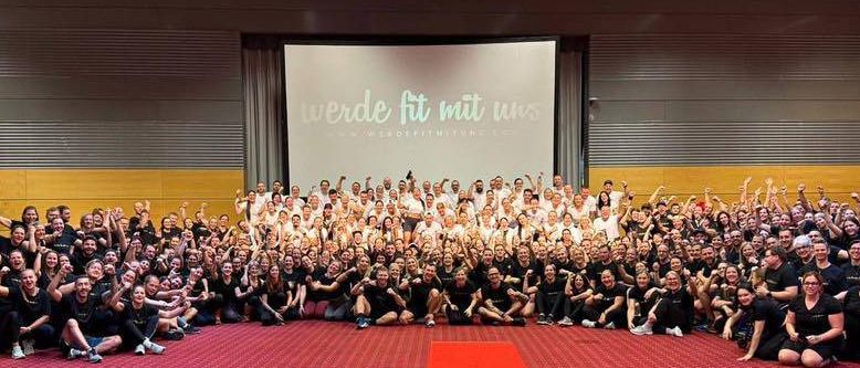 Eine Gruppe von Menschen in passender schwarzer Kleidung posiert für ein Foto in einem Raum mit rotem Teppich und Beamerleinwand.
