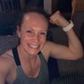 Eine Frau lächelt und reckt ihren Arm, trägt ein graues Tanktop und eine weiße Armbanduhr. Ihr Haar ist zerzaust, und sie scheint sich in einem dunklen Raum zu befinden.