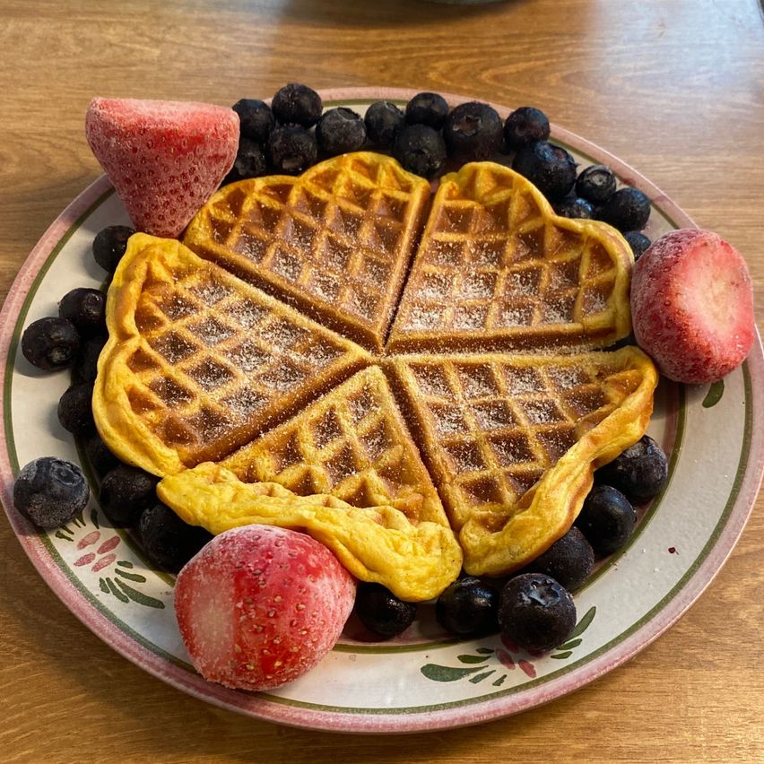 Ein Teller mit Waffeln, Erdbeeren und Blaubeeren. Die Waffeln sind knusprig, goldbraun und mit Zucker bestreut. Im Hintergrund befindet sich ein Holztisch.