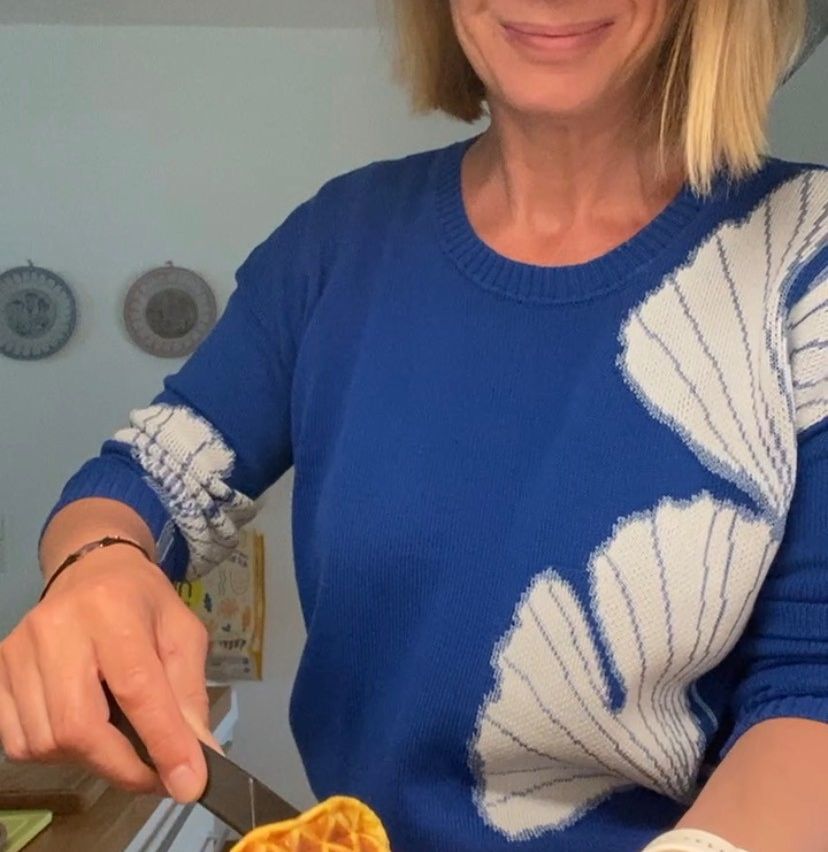 Eine Frau mit blondem Haar schneidet eine Orange mit einem Messer. Sie trägt einen blauen Pullover mit einem weißen Blattmuster.
