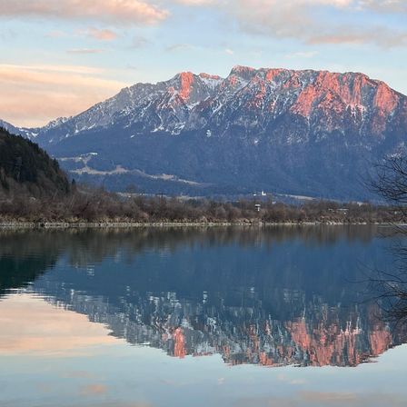 Eine ruhige Berglandschaft mit einem ruhigen See, der den rötlichen Schimmer der Berge bei bewölktem Himmel widerspiegelt.