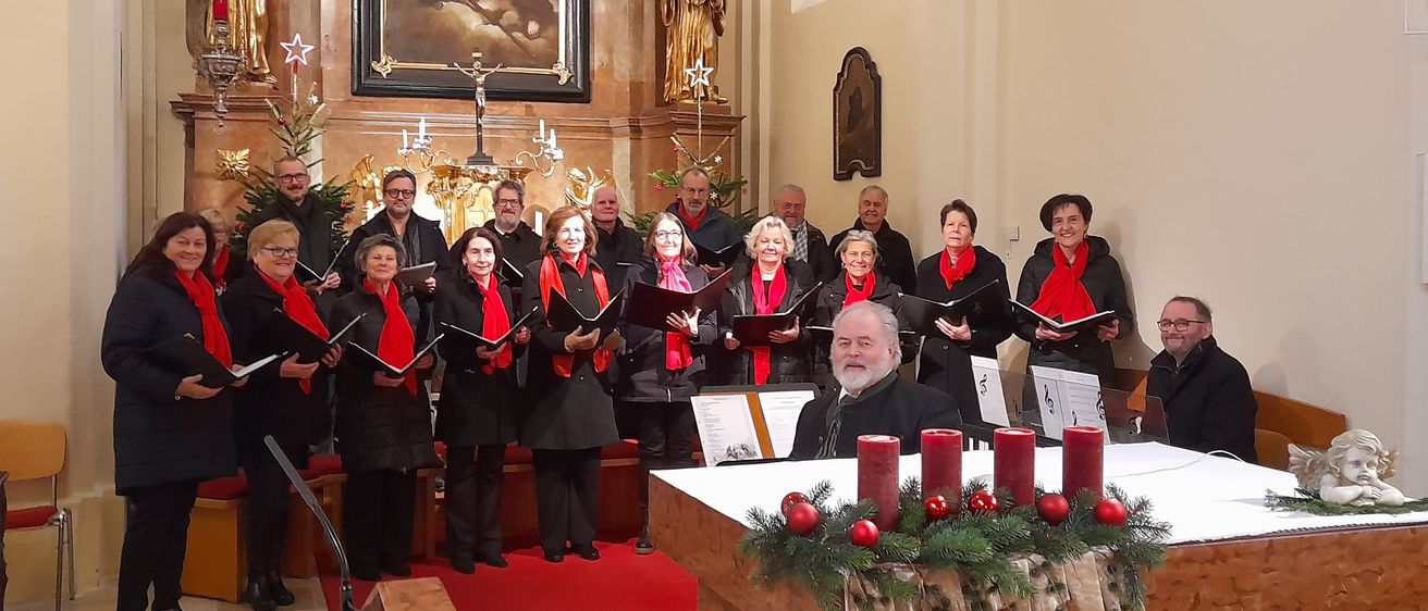 Ein Chor von Erwachsenen, gekleidet in rote Schals und schwarze Mäntel, steht in einer Kirche, hält Notenblätter und singt. Eine goldene Statue und ein Gemälde schmücken die Wände.