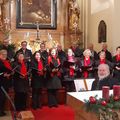 Ein Chor in roten Schals steht in einer Kirche, hält Bücher in der Hand, mit einer goldenen Statue dahinter. Auf dem Altar stehen drei Kerzen, und eine Weihnachtsbaum ist links.
