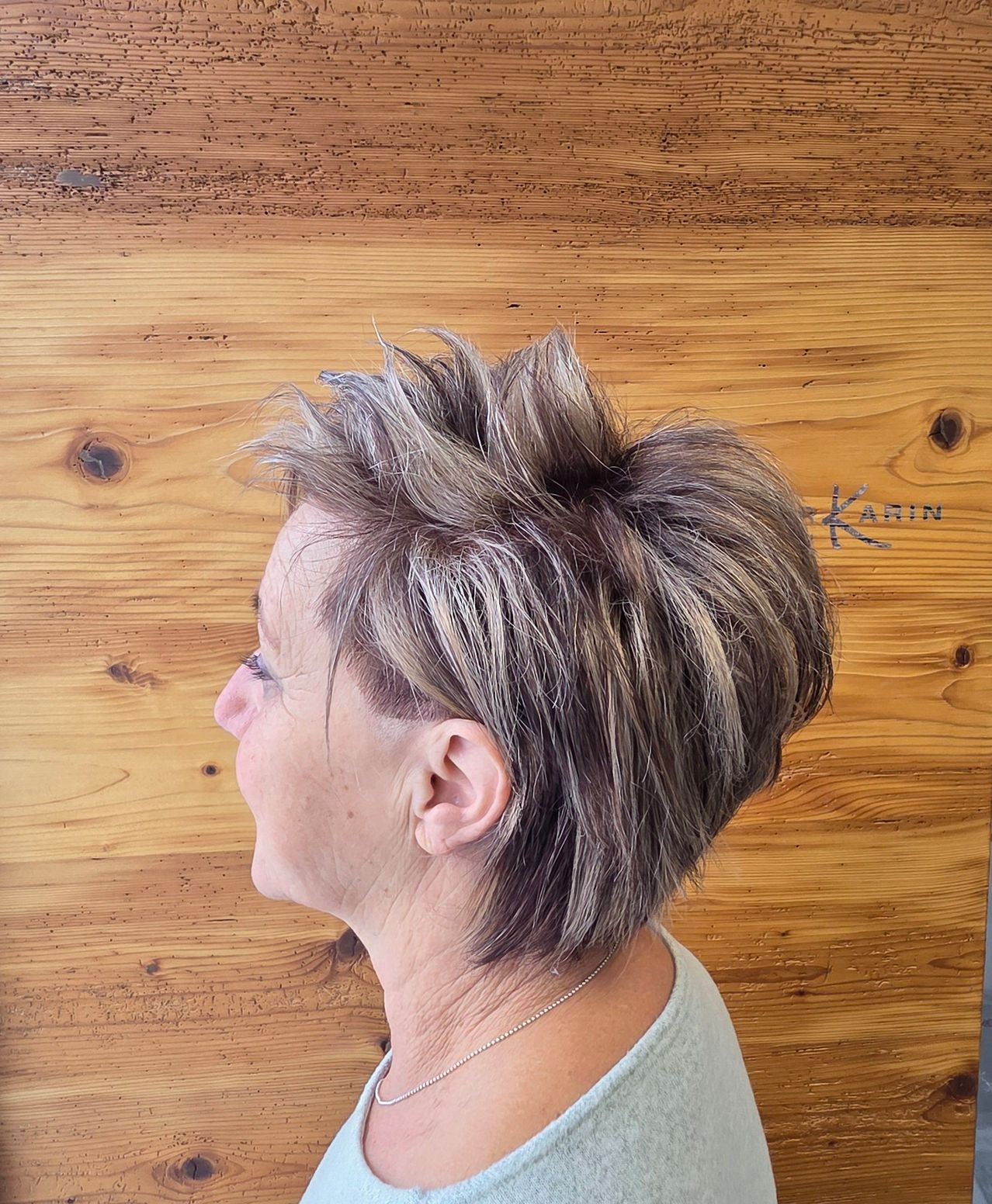 Eine Frau mit kurzen, blonden Haaren, die in einem Irokesenschnitt gestylt sind, schaut nach links. Sie trägt eine Halskette. Hinter ihr befindet sich eine Holzwand mit sichtbaren Knoten und Maserungen.