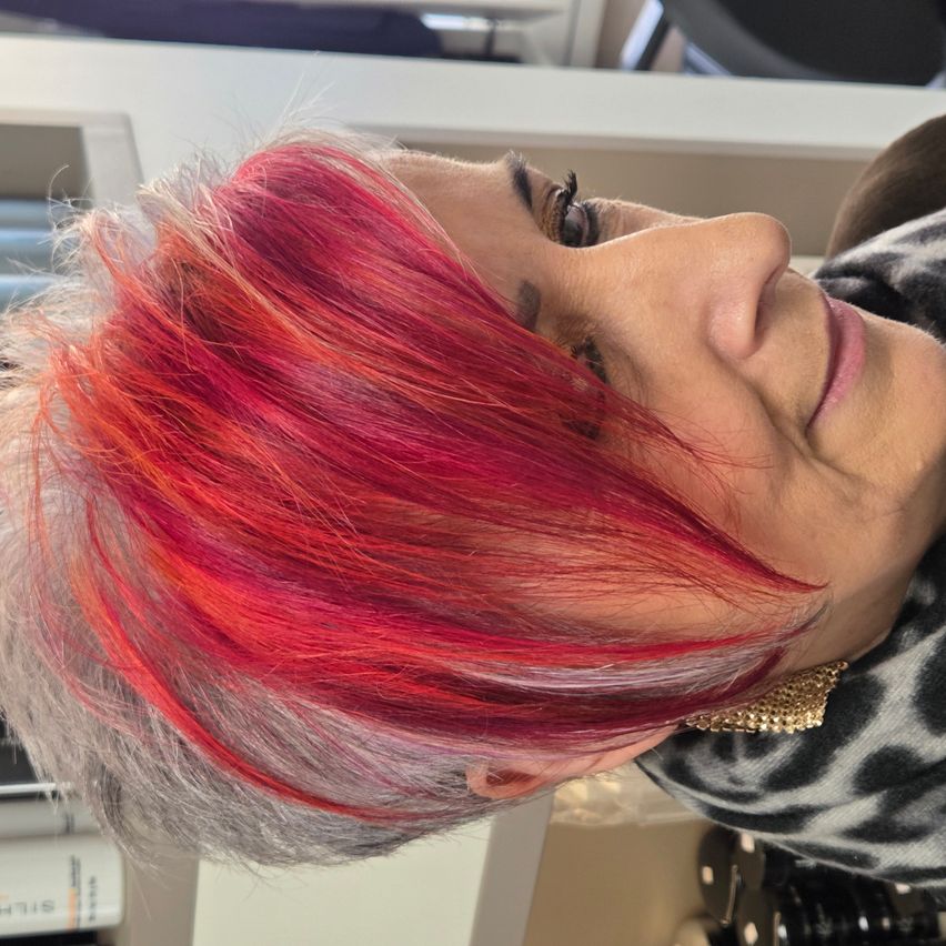 Eine Frau mit einem Mix aus rotem und grauem Haar blickt mit einem leichten Lächeln nach vorne. Sie trägt einen Leopardenmuster-Schal und goldene Ohrringe.