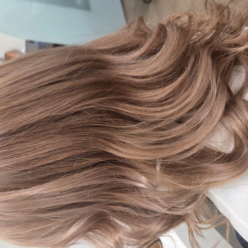 Nahaufnahme von langen, welligen, hellbraunen Haaren, die über eine weiße Oberfläche fallen. Das Haar hat einen natürlichen Glanz und sieht gesund aus.