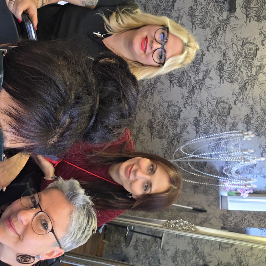 Vier Frauen posieren für ein Selfie, zwei mit Brille, eine mit Halskette und rotem Lippenstift und eine weitere mit Armband. Der Hintergrund hat eine verzierte Tapete.