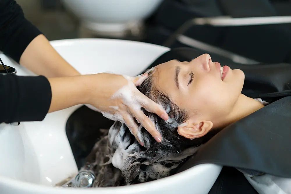 Eine Frau mit geschlossenen Augen lässt sich in einem Salon die Haare waschen. Die Person, die ihr die Haare wäscht, trägt ein schwarzes Langarmhemd.