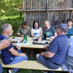 Eine Gruppe von Menschen sitzt um einen Holztisch im Freien, lächelt und lacht. Sie genießen Essen und Getränke. Der Tisch ist mit Sandwiches, Getränken und Servietten gedeckt. Hinter ihnen steht ein Holzzaun.