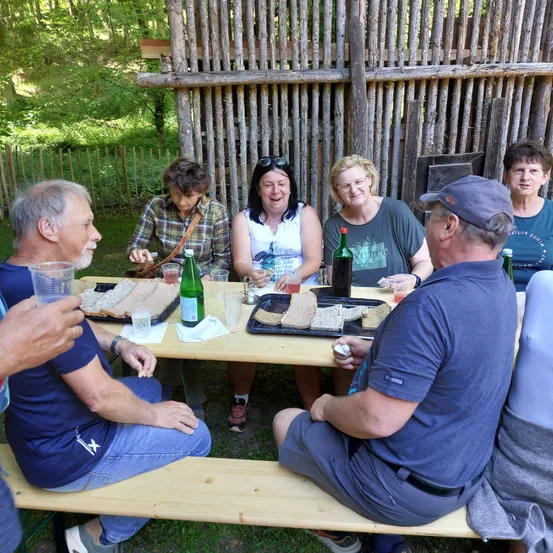 Eine Gruppe von Menschen sitzt um einen Holztisch im Freien, lächelt und lacht. Sie genießen Essen und Getränke. Der Tisch ist mit Sandwiches, Getränken und Servietten gedeckt. Hinter ihnen steht ein Holzzaun.